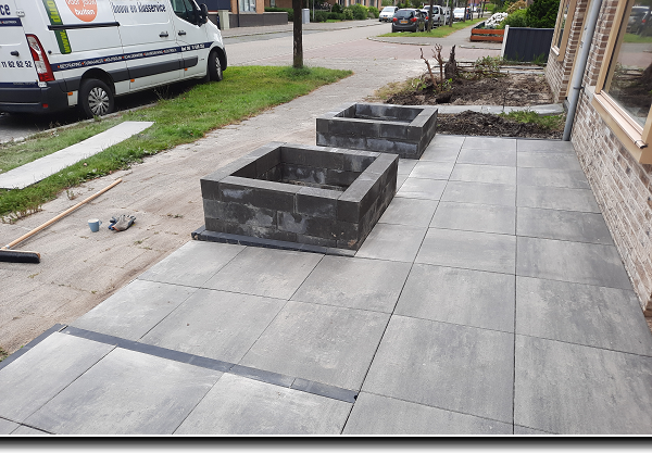 Bestrating van grote grijs-antraciet kleurige tegels met twee bloembakken van zwarte lineablokken